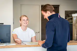 Zwei Personen an einer Rezeption im Campingbereich, eine spricht, die andere schaut auf den Schalter, im Hintergrund Türen und Fenster.