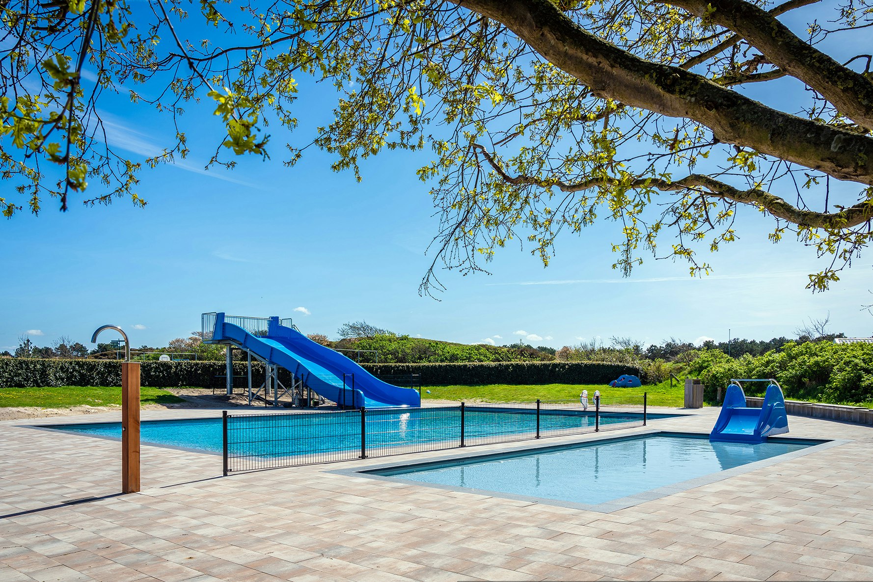 Schwimmbecken mit blauer Rutsche und Treppe, umgeben von Holzterrasse und Hecken, im Hintergrund Wiese und Bäume.