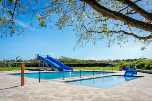 Schwimmbecken mit blauer Rutsche und Treppe, umgeben von Holzterrasse und Hecken, im Hintergrund Wiese und Bäume.