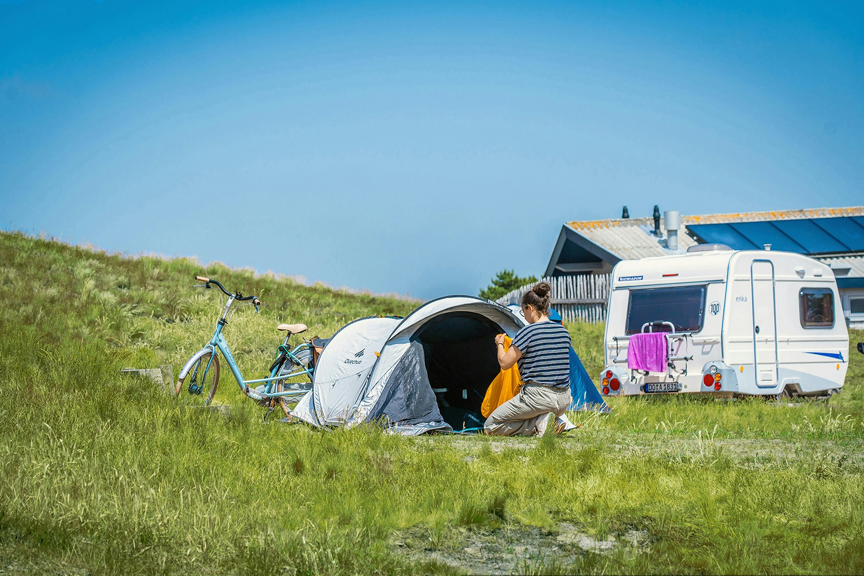 Campingzelt und Fahrrad auf einer Wiese, daneben ein Wohnmobil; zwei Personen sitzen am Eingang des Zelts.