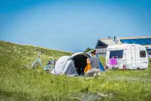 Campingzelt und Fahrrad auf einer Wiese, daneben ein Wohnmobil; zwei Personen sitzen am Eingang des Zelts.