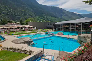 Großes Schwimmbecken mit Sonnenschirmen und Liegen, daneben ein Glasdach-Pavillon, im Hintergrund Berge und Campinganlage.