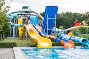 Bunte Wasserrutschen am Camping-Wasserpark, Poolbecken im Vordergrund, Rutschen und Plattformen vor grünen Bäumen.