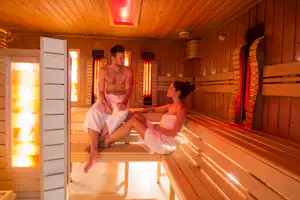 Sauna im Innenraum mit Holzverkleidung, Bankreihen und roten Lichtsäulen, zwei Personen sitzen in Handtüchern.