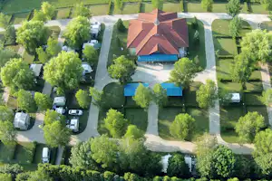 Luftaufnahme: Gebäude mit rotem Dach, Stellplätze mit Wohnmobilen und Wohnwagen, Wege und Hecken im grünen Campinggelände.