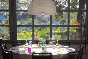 Runder Tisch im Camping-Restaurant mit Gläsern und Karaffen, große Fensterfront mit Blick auf Bäume und Garten.