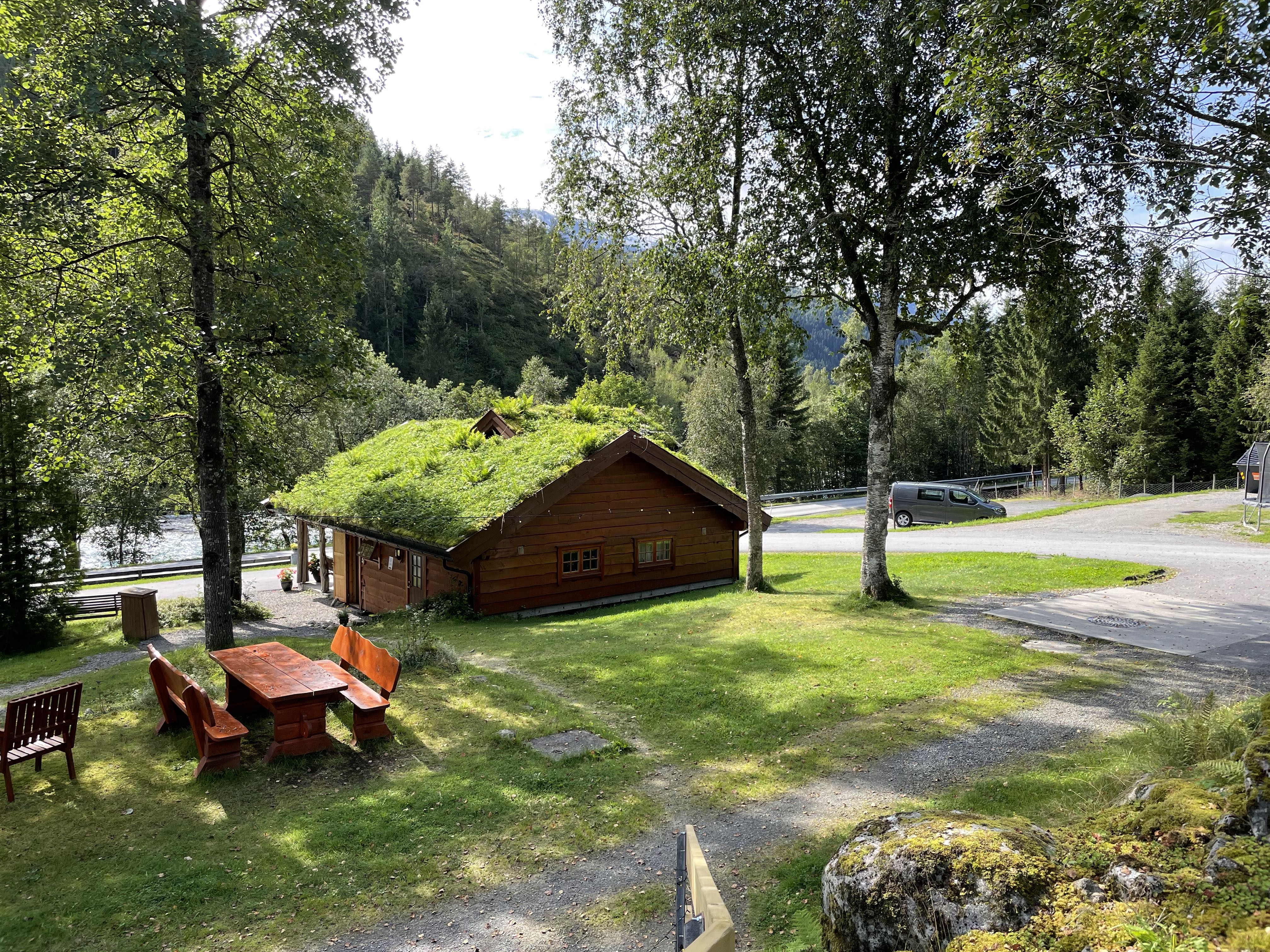 Holzhütte auf einem Campingplatz im Grünen, mit Sitzgruppe und Weg, umgeben von Bäumen und Wiese.