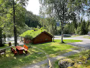 Holzhütte auf einem Campingplatz im Grünen, mit Sitzgruppe und Weg, umgeben von Bäumen und Wiese.