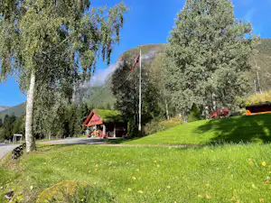 Grüne Campingwiese mit Bäumen, Flaggenmast und kleinen Gebäuden am Hang, im Hintergrund ein Berg.