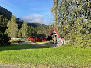 Rote Hütte am Campingplatz mit Veranda, Rasenfläche und Weg, umgeben von Bäumen und Bergen im Hintergrund.