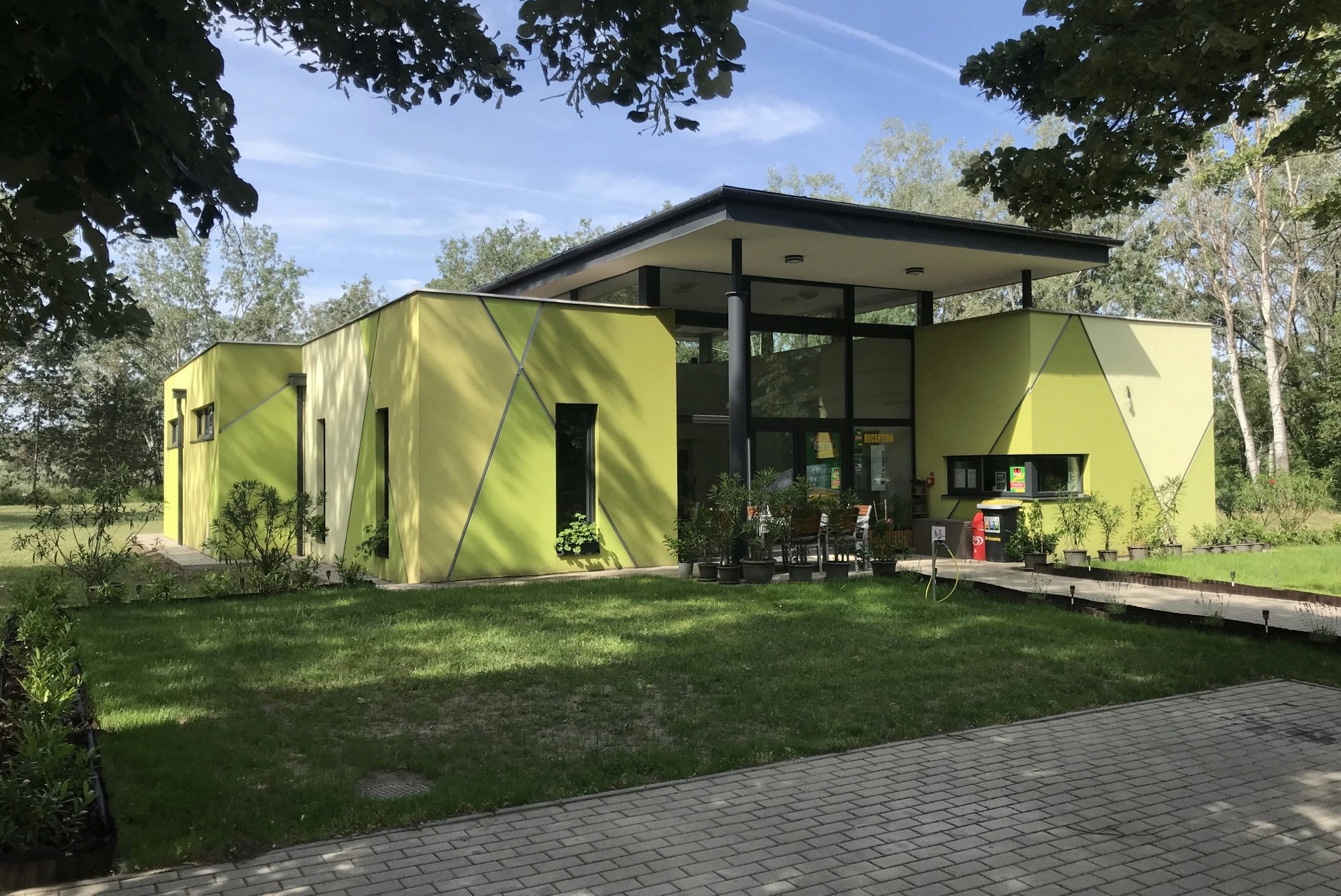 Grünes Ferienhaus mit überdachter Terrasse, Sitzgruppe und Tisch auf dem Campingplatz, umgeben von Rasen und Bäumen.