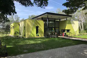 Grünes Ferienhaus mit überdachter Terrasse, Sitzgruppe und Tisch auf dem Campingplatz, umgeben von Rasen und Bäumen.