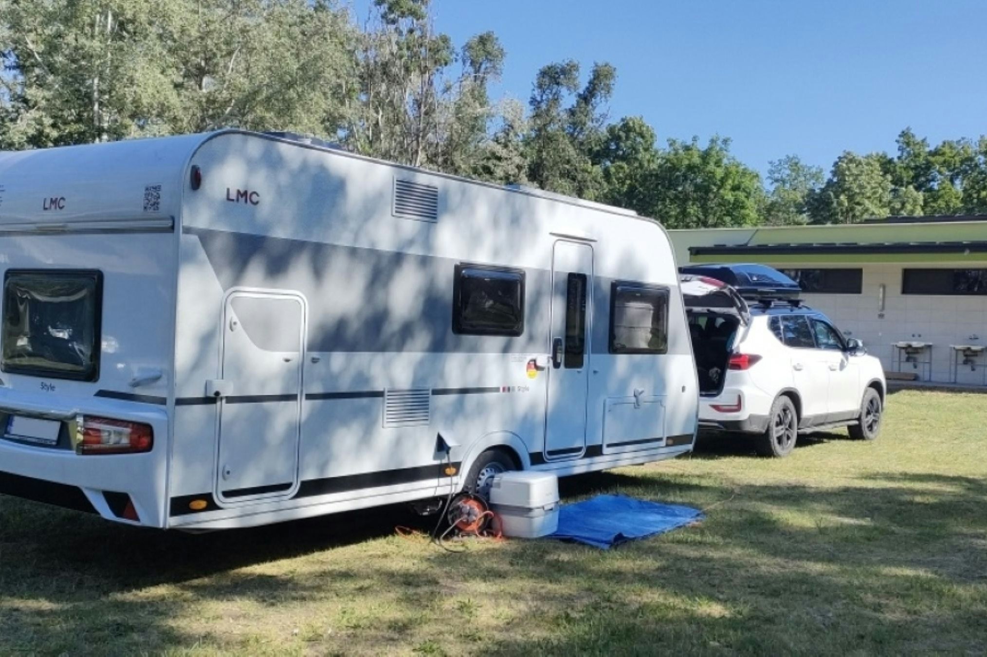 Wohnwagen auf einem Campingplatz-Stellplatz, daneben ein Auto und ein Gebäude mit grüner Fassade im Hintergrund.