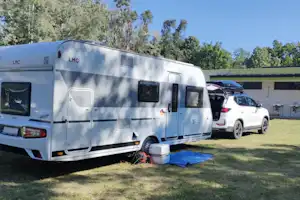 Wohnwagen auf einem Campingplatz-Stellplatz, daneben ein Auto und ein Gebäude mit grüner Fassade im Hintergrund.