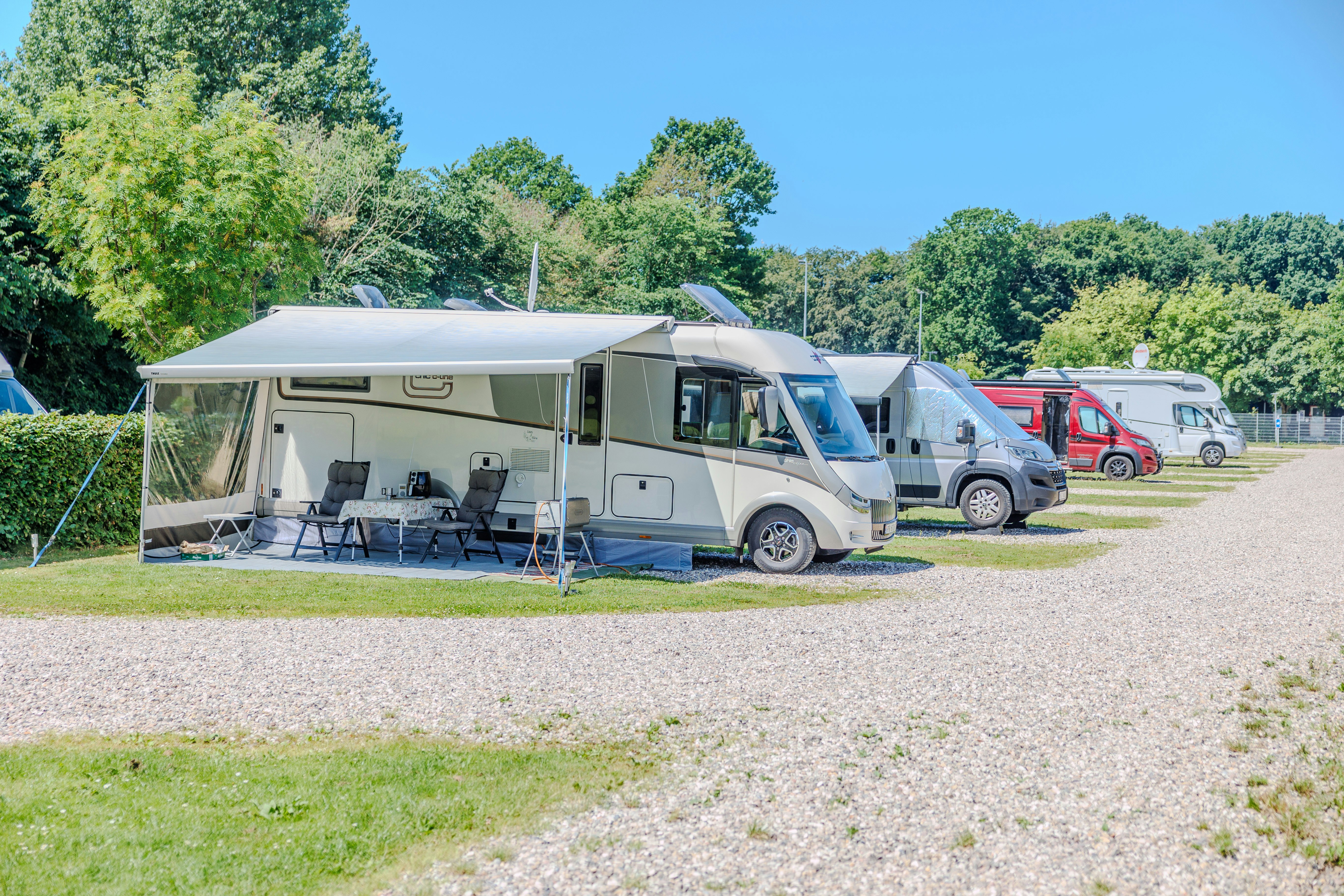 Wohnmobil auf dem Campingplatz mit Vorzelt, Tisch und Stühlen, daneben weitere Fahrzeuge und Bäume im Hintergrund.