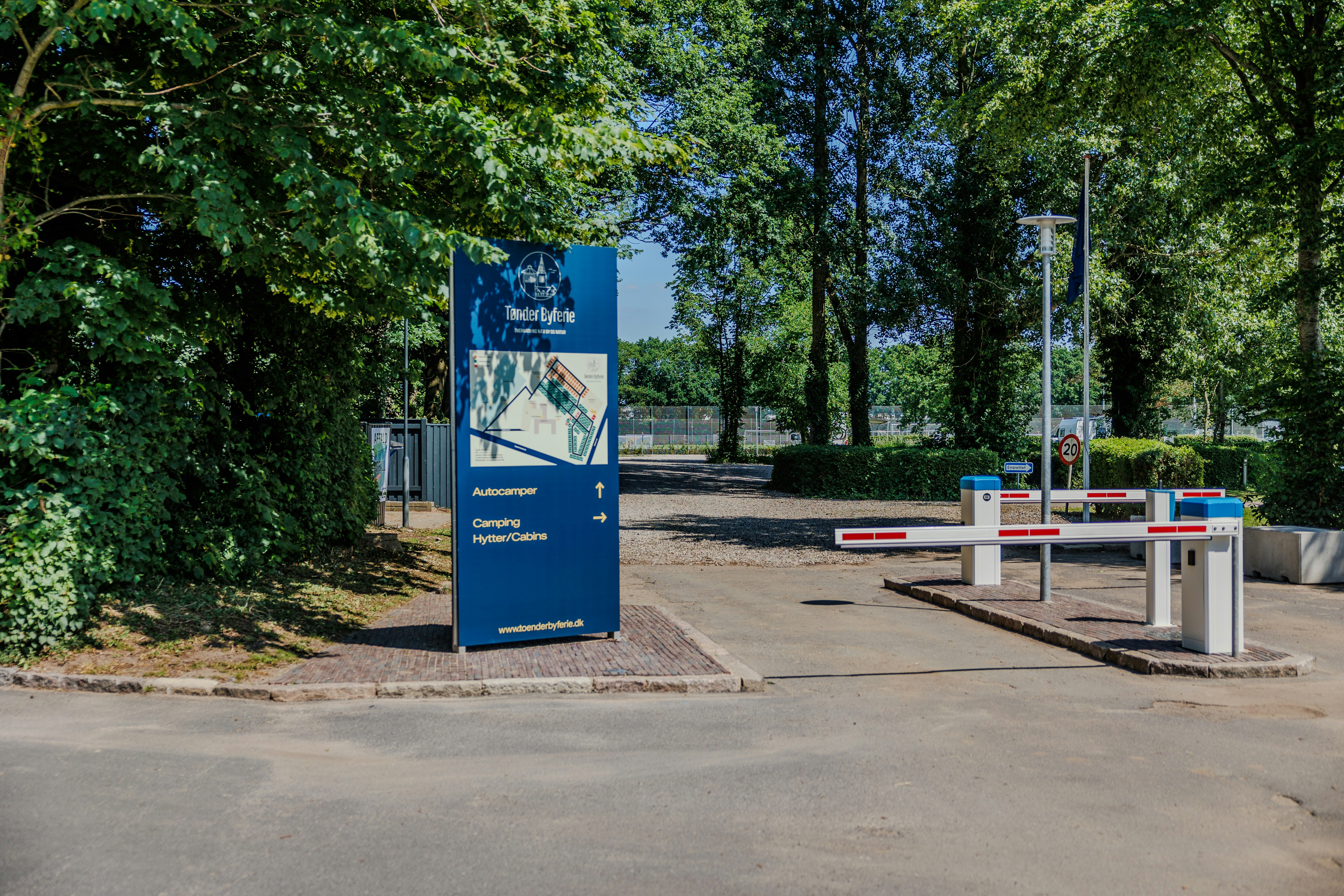 Eingangsschild am Campingplatz neben dem Parkplatz, umgeben von Bäumen und Hecken, mit Schrankenanlage im Hintergrund.