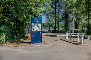 Eingangsschild am Campingplatz neben dem Parkplatz, umgeben von Bäumen und Hecken, mit Schrankenanlage im Hintergrund.