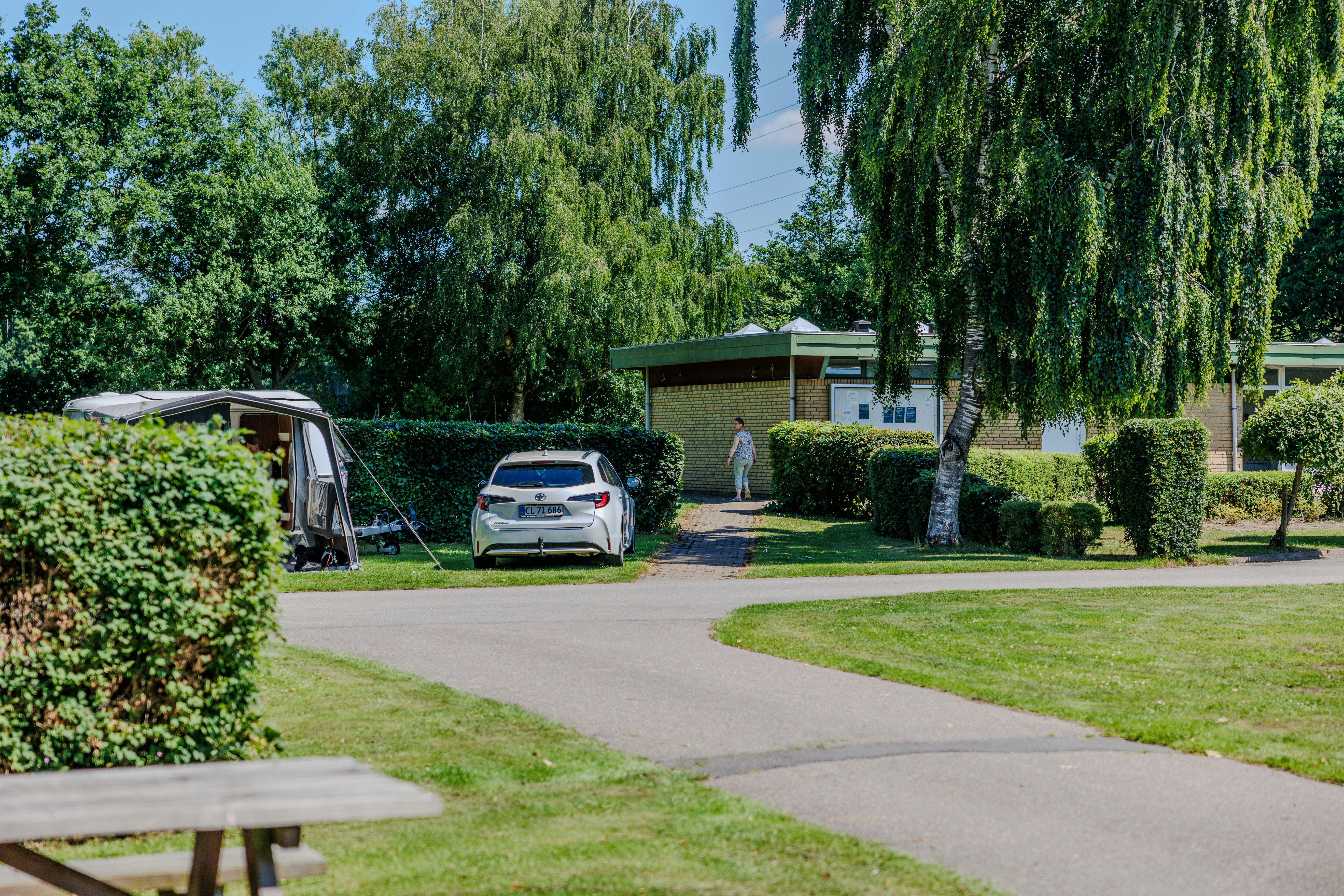 Wohnwagen auf einem Campingplatz-Stellplatz, gepflasterter Weg, Hecken und ein Baum im Vordergrund.