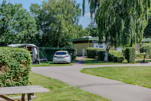 Wohnwagen auf einem Campingplatz-Stellplatz, gepflasterter Weg, Hecken und ein Baum im Vordergrund.