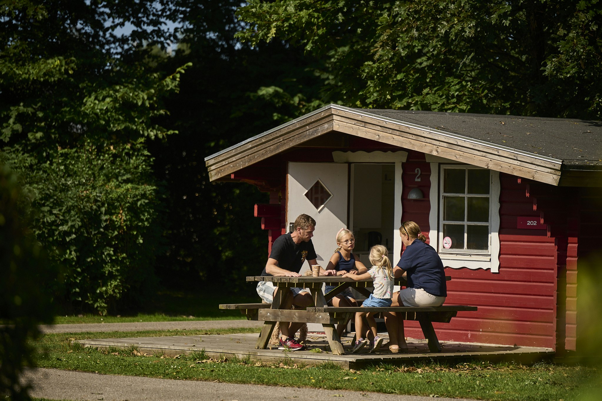 Holz-Hütte mit roter Fassade und Veranda, davor Tisch und Bänke; mehrere Personen sitzen und essen im Grünen.