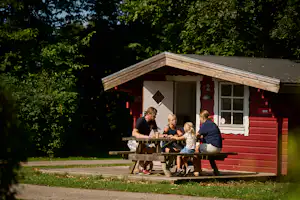 Holz-Hütte mit roter Fassade und Veranda, davor Tisch und Bänke; mehrere Personen sitzen und essen im Grünen.