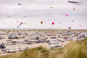 Campingplatz am Strand mit vielen Wohnwagen und Zelten, im Himmel fliegende Drachen über dem Meer.