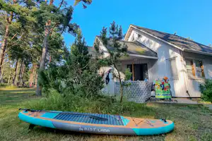 Vor einem weißen Campinghaus steht ein blaues SUP-Board auf dem Rasen, umgeben von Bäumen und einem kleinen Sitzbereich.