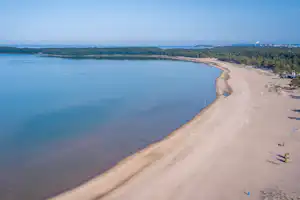 Weites Seeufer mit Sandstrand und Bucht, Blick von oben auf Wasser, Uferlinie und einzelne Liegen am Rand.