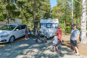 Wohnwagen auf dem Campingplatz im Wald, daneben Auto und Familie mit Kind auf dem Weg.