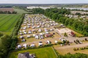 Luftaufnahme eines Campingplatzes mit vielen Wohnmobilen und Wohnwagen, Wegen, Bäumen und einem großen See im Hintergrund.