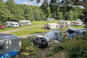 Campingplatz mit Wohnwagen und Zelten auf einer Rasenfläche, Auto und Anhänger im Stellbereich, umgeben von Bäumen.