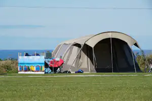 Großes Campingzelt am Meer auf einer grünen Wiese, daneben Stühle und Tisch, im Hintergrund Wasser und Küste.