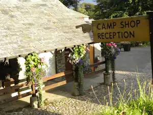 Holzschild „CAMP SHOP RECEPTION“ am Eingang neben Steinwand, Blumenampeln und Holzgeländer am Campingplatz.