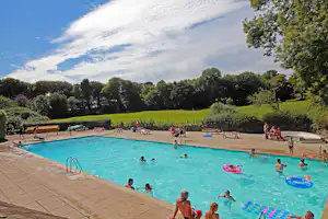 Großes Camping-Schwimmbecken mit Liegen, Menschen im Wasser und bunten Schwimmreifen, umgeben von Grün und Bäumen.