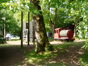 Campingplatz im Schatten großer Bäume mit rotem Gerät, Schildern und einem Weg im Hintergrund.