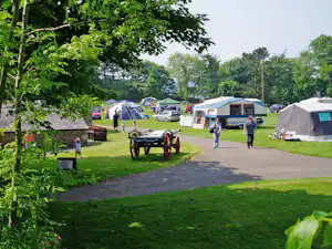 Campingplatz mit Zelten und Wohnwagen, breiter Weg zwischen den Stellplätzen, Personen gehen vorbei, Bäume und Rasen im Vordergrund.