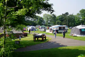 Campingplatz mit mehreren Zelten und Wohnwagen, Rasenflächen, breitem Weg und Bäumen im Hintergrund.
