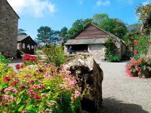 Stein-Hütte mit Holzfront und Terrasse, Kiesweg und Blumenbeete mit bunten Blüten im Campingbereich.