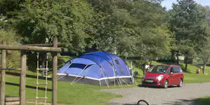 Blaues Zelt auf einem Campingplatz mit rotem Auto daneben, Grasweg und Bäume im Hintergrund.