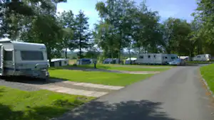 Wohnwagen auf einem Campingplatz, gepflasterte Wege und Rasenflächen zwischen Stellplätzen, im Hintergrund weitere Fahrzeuge und Bäume.