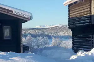 Zwei Campinghütten aus Holz im Schnee, dazwischen ein Weg, Blick auf verschneite Berge und Bäume im Winter.