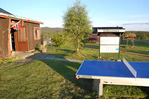Tischtennisplatte auf dem Campingplatz, daneben Rasen, Bäume und ein Holzgebäude mit Eingang.