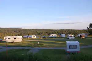 Campingwiese mit Wohnwagen und kleinen Gebäuden, gepflasterter Weg und Bäume am Horizont.