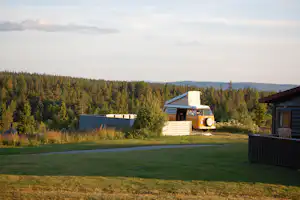 Wohnmobil auf einem Campingplatz mit Rasenfläche, Hecke und Bäumen im Hintergrund, daneben ein kleines Gebäude.