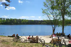 Fahrräder am Ufer eines Sees, daneben Sitzgelegenheiten und Tische auf einem Kiesstrand, Bäume und Wald im Hintergrund.