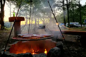 Lagerfeuer mit Funken und Grillrost über dem offenen Feuer, umgeben von Bäumen und Campingfahrzeugen am Rand.
