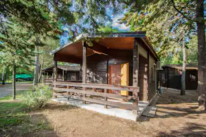 Holzhütte mit Veranda und Treppe am Campingplatz im Wald, umgeben von Bäumen und einem Kiesweg.