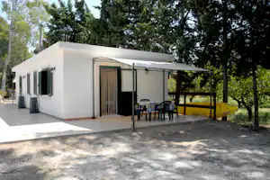 Weißes Ferienhaus mit Terrasse, Tisch und Stühlen vor hohen Bäumen im Campingbereich.