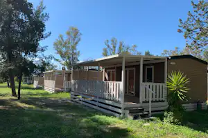 Bungalow mit Veranda und Holzgeländer auf einem grünen Campingplatz, umgeben von Bäumen und Rasen.