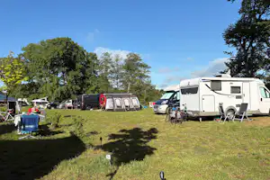 Wohnwagen und Zelte auf einem Campingplatz, daneben ein Auto und ein Wohnmobil, Rasenfläche mit Bäumen im Hintergrund.